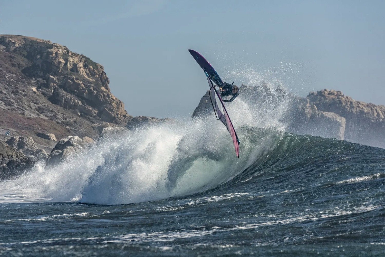 A windsurfer in the air above a wave below the coastal hills